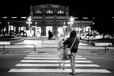 Rear view of man with shopping cart walking on zebra crossing at night