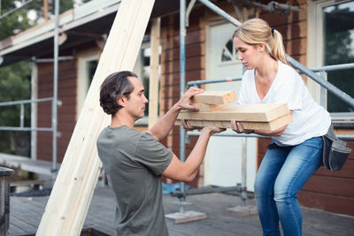 Side view of couple carrying stack of lumber outside house being renovated