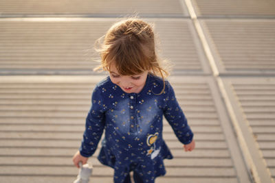 High angle of adorable cheerful little girl in blue dress with toy on hand having fun on paved walkway in summer day