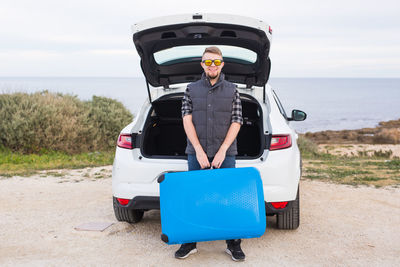 Portrait of man standing by car on sea shore against sky