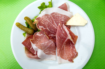 Directly above close-up of ham with pickles and cheese served in white plate against green background