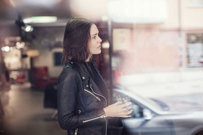 Side view of woman wearing black leather jacket holding drink while standing in workshop seen from window