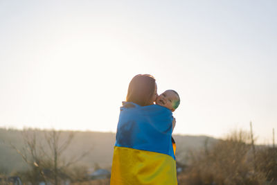 Woman hugs her little son wrapped in yellow and blue flag of ukraine in outdoors.