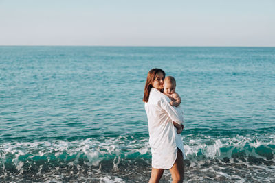 A young mother walks along the beach with a small child in diapers.