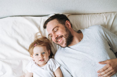 Young happy family with father and daughter baby girl on bed in cozy home, view from top