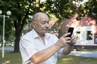 An elderly man takes pictures on a smartphone camera in a park outdoors.