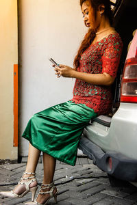 Side view of woman using smart phone while sitting on car trunk