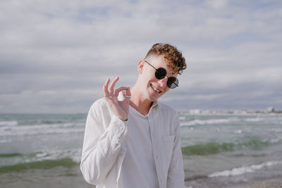 Young guy in braces with round sunglasses at the sea shows okay