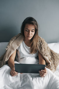 Sick young woman using digital tablet on bed against wall at home