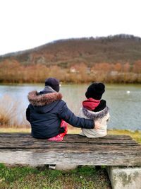 Rear view of siblings sitting on wooden bench by lake