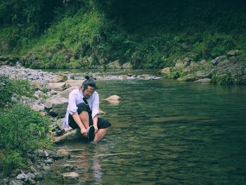 Man removing shoe while sitting on rock by stream