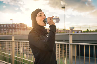 Beautiful woman wearing hijab drinking water against building during sunrise