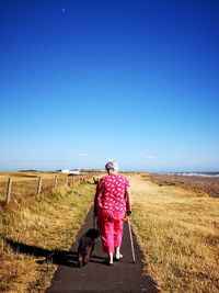 Full length rear view of woman with dog walking on road against clear sky during sunny day