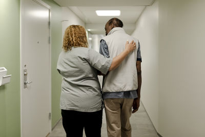Rear view of female nurse walking with arm around senior man in corridor at retirement home