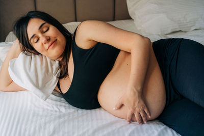 Middle-aged brunette pregnant woman hugging her tummy at home while lying on bed