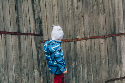 A child in a blue jacket and a hat walks near a wooden wall. real and serene people