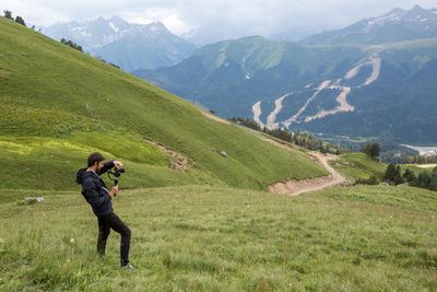 Guy kameraman in a jacket stands in the mountains on a green meadow and shoots a video on steadicam