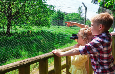 Side view of boy gesturing while standing by girl looking through binoculars