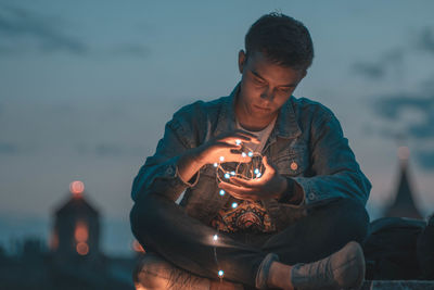 Man with illuminated string light sitting against sky