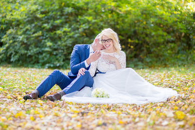 Portrait of smiling bride and groom sitting on field against plants at park