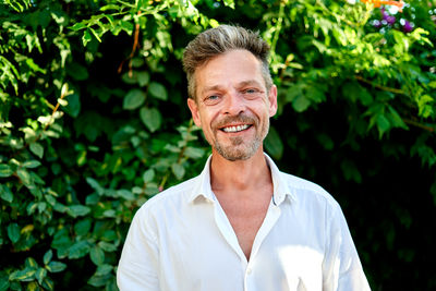 Happy middle aged businessman in white shirt clasping hands and looking at camera while standing near lush bushes on summer day in garden