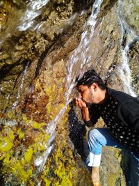Young man drinking water from waterfall against rocks