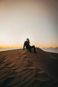 Man sitting on sand in desert against sky during sunrise