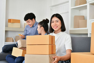 Portrait of smiling businesswoman sitting with boxes while colleagues using phone at office