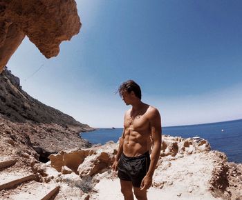 Full length of shirtless man standing on rock at beach against sky