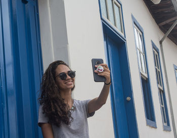 Smiling young woman taking selfie while standing by wall