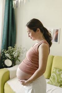 Pregnant woman holding abdomen standing by sofa at home