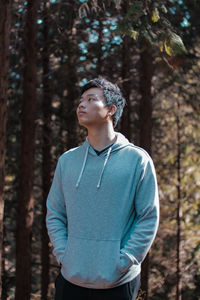 Young man looking away while standing against trees