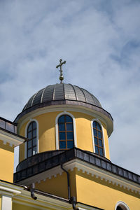 View from below of church tower with arched windows and metal dome with cross against cloudy sky.