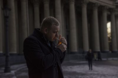 Young man igniting cigarette while standing against columns during winter