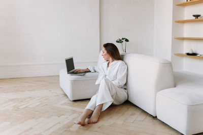 A millennial woman in white pajamas looks into a mobile phone and works online using technology