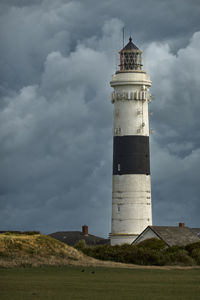 White lighthouse with a black stripe behind a meadow and houses in front of a dramatic cloudy sky