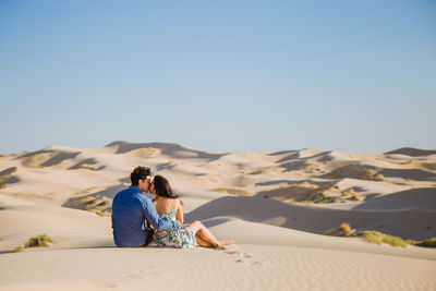 Rear view of couple kissing while sitting on sand at desert against sky