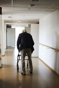 Full length rear view of senior man walking with rollator in hospital corridor