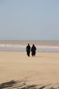 Rear view of female friends walking at beach against clear sky