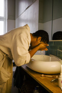 Side view of young woman washing face at sink in bathroom