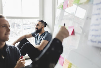 Businessman sitting on window with colleagues looking at whiteboard in foreground