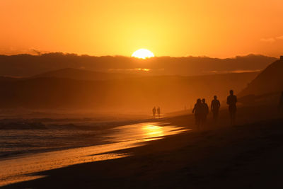 Silhouette people walking on beach against orange sky