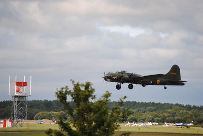 Military airplane flying over landscape against cloudy sky