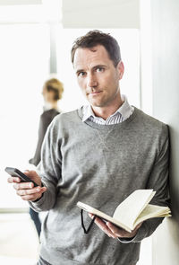 Portrait of mid adult businessman with mobile phone at book standing in office