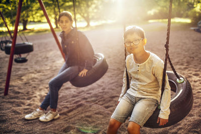 Portrait of brother and sister sitting on swing at park during sunny day