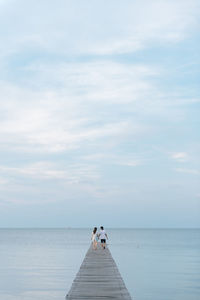 Rear view of couple walking on pier over sea against cloudy sky