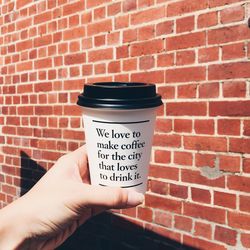 Cropped hand of woman holding disposable cup against brick wall