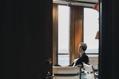 Thoughtful businesswoman looking through window sitting in board room seen from doorway