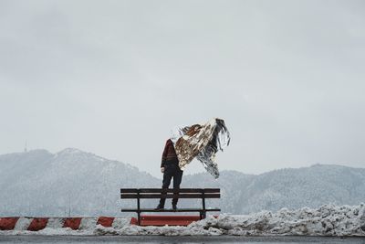 Man holding foil while standing on bench against mountain and sky during winter