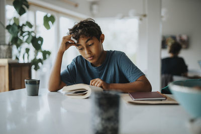 Boy leaning on elbow while reading book near table at home